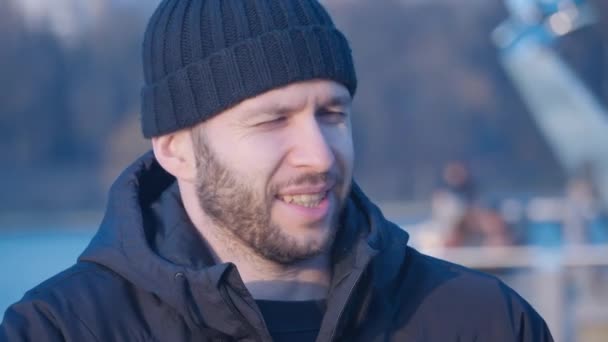 Porträt eines jungen, gutaussehenden, bärtigen Mannes, der vor Kälte zitternd eine schwarze Jacke zuknöpft, lächelt und in die Sonnenuntergangssonne auf dem Hintergrund eines blauen Sees im zentralen Stadtpark blickt. — Stockvideo