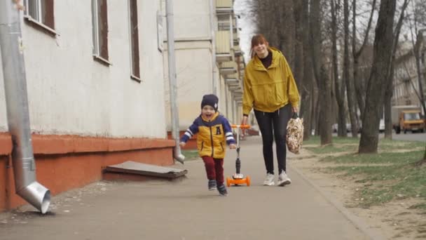 Menino feliz com um sorriso encantador corre pela estrada para conhecer o pai, atrás dele a mãe rola uma scooter. Movimento lento . — Vídeo de Stock