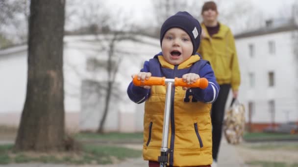 Charmante kleine jongen rijden op een scooter. Op de achtergrond loopt een vrouw de Defocus. Slow motion-video — Stockvideo