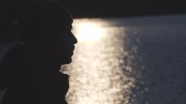 yavaş hareket merkezi şehir parkında mavi bir gölde güneş gün batımı güneş yansıması arka planda Genç sakallı bir adamın silüeti 4k video yakın.