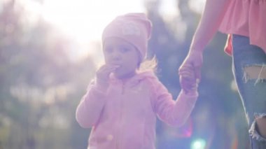 Genç bir kadın ve küçük kızı gün batımında parkta yürüyorlar ve parlak güneş onlara parlıyor ve annesi de kızların elini tutuyor. Mutlu aile hayatı konsepti 4k ağır çekim videosu