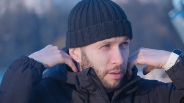 siyah ceketinin bir kaputunu giyen ve merkez şehir parkında mavi bir gölün arka planında gün batımı güneşine bakan genç yakışıklı sakallı bir adamın portresi 4k videoya yakın.