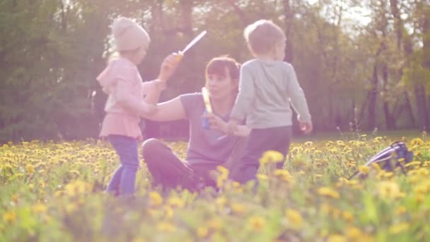 Piękna kobieta uczy jej małego syna i córki cios pęcherzyki mydła w parku miejskim na jasnym słoneczny dzień wiosny. Szczęśliwa matka i jej dzieci są na wakacjach koncepcji w zwolnionym tempie 4K wideo — Wideo stockowe