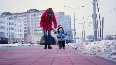 Genç bir kadın kışın sokağın aşağısında bir bebeğe yol gösteriyor. İlk adımlar bebeğim.