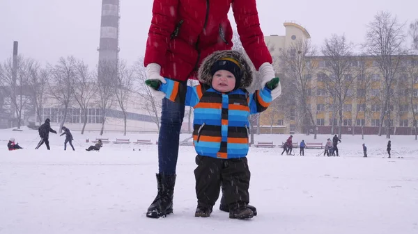La mamma guida le mani di un piccolo bambino sorridente nella neve del parco. I primi passi del bambino. Assistenza parentale — Foto Stock