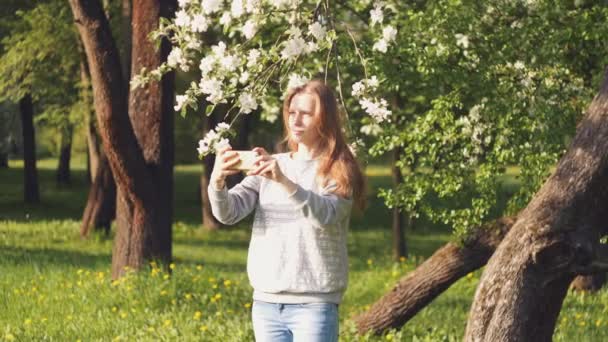 Çiçek açan bir ağacın arka planındaki güzel beyaz kız selfie çeker, saçını düzleştirir. — Stok video