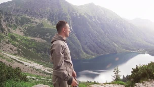 Turista no topo de uma montanha fotografa um lago de montanha. Dia ensolarado. Bela vista do lago da montanha. O conceito de liberdade, felicidade e aventura. Vídeo 4K — Vídeo de Stock