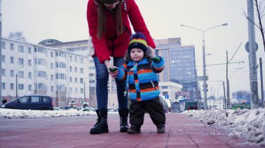 Genç bir kadın kışın sokağın aşağısında bir bebeğe yol gösteriyor. İlk adımlar bebeğim.