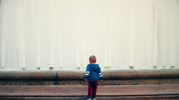 小さな男の子が広場に立っていて、大きく美しい噴水を見ています。. — ストック写真
