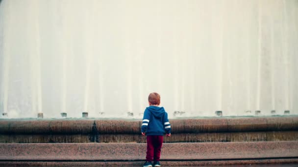 Un niño pequeño se para en la plaza y mira una fuente grande y hermosa. — Vídeo de stock