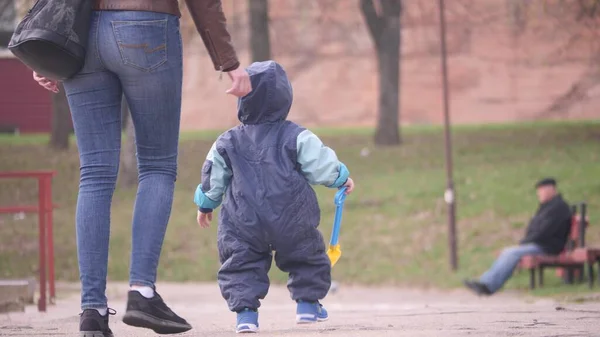 赤ちゃんのシャベルを持っている彼女の小さな息子と若い女性は、 4Kで春の曇りの日に市内公園の池の近くを歩いていますバックビュースローモーションビデオ. — ストック写真