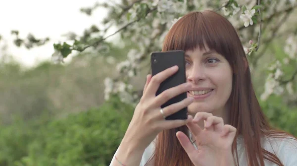 Kız bahçede selfie çekiyor. Çekici kızıl saçlı bir kadın kiraz bahçesinde cep telefonu kullanarak selfi yaparken gülümsüyor. Cihazları sağlıklı bir yaşam tarzı için kullanma kavramı.