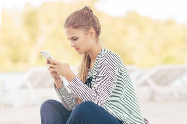 Sahilde bir akıllı telefon olan kadın. Tatil ve teknoloji. Telefonunuzun ve açık havada deniz kenarında bir plajda oturmuş rahat kıyafetler kız şirin şık flowned. Yakın çekim portre çekici