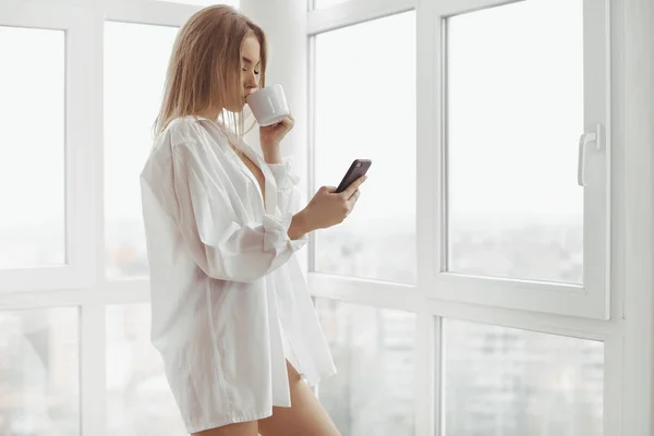 Prachtige blond meisje met smartphone en kopje koffie binnenshuis. — Stockfoto