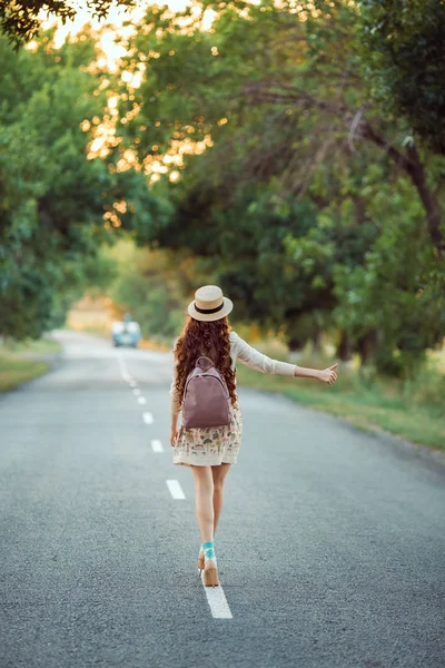 Девушка в шляпе и рюкзаке автостопом по дороге — стоковое фото