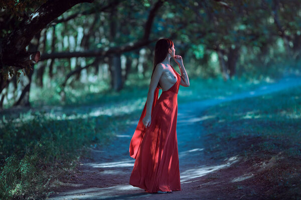 Wonderful sexy fashion model with naked walking in a fantastical forest. Photo of seductive nude woman in full length in luxury long red dress posing against bokeh trees background. Multi-racial Asian