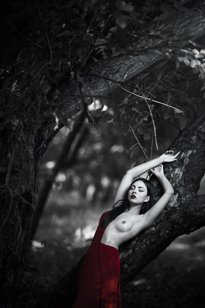 Sensual sexy fashion model with naked breast resting in fantastical forest. Photo of seductive nude woman in luxury long red dress laying on tree. Multi-racial Asian Caucasian girl. Fashionable toning