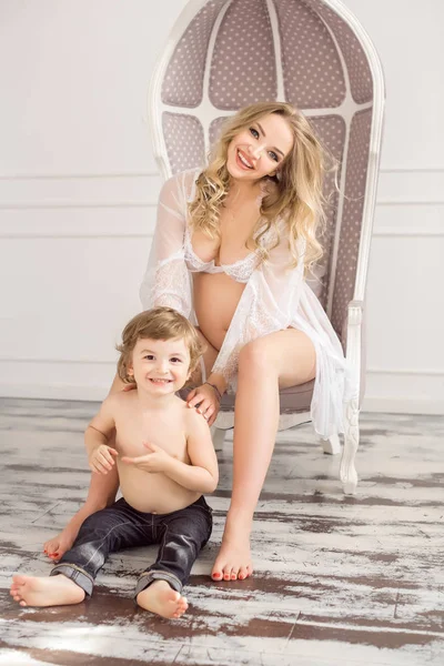 Blonde pregnant woman with her child boy in white indoor on chair — Stock Photo, Image