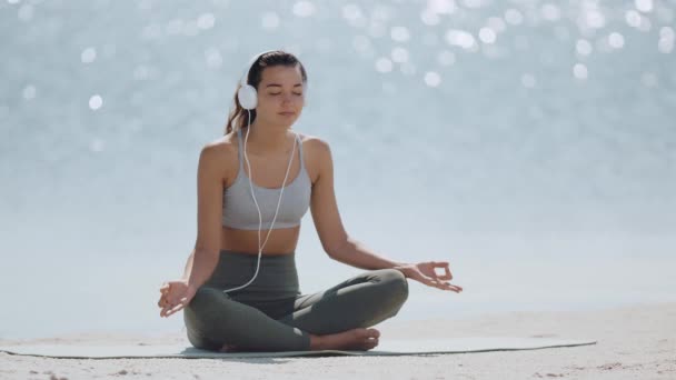 Yoga yapan kadın meditasyon yapıyor Sahilde kulaklıkla poz veriyor — Stok video