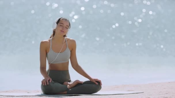 Женщина в позе медитации йоги с наушниками на пляже — стоковое видео