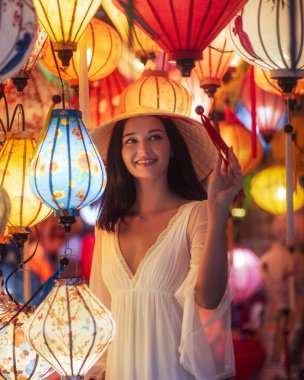 Geleneksel Vietnam şapkalı ve zarif beyaz elbiseli çekici Asyalı kadın Hoi An 'da renkli fenerlerle çevriliyken gülümsüyor. Moda, turizm ve kültür görselleri için ideal..
