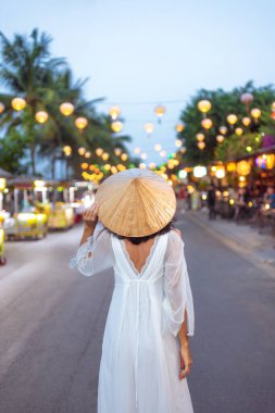 Uçan beyaz elbiseli ve konik şapkalı bir kadının Hoi An, Vietnam 'da fenerle aydınlatılmış bir caddede akşam karanlığında yürüyüşü. Seyahat reklamları, yaşam tarzı blogları ve kültürel kampanyalar için ideal..
