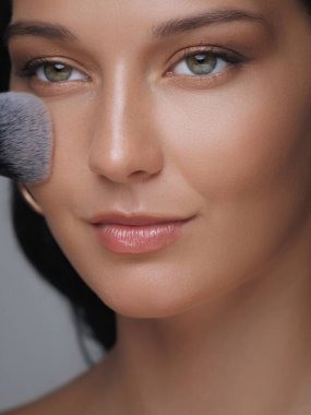 Pudra sürmek için makyaj fırçası kullanan parlak ciltli güzel bir genç kadın. Güzellik konseptleri, üst düzey kozmetik markalaşması ve kişisel bakım promosyonları için mükemmel.. 
