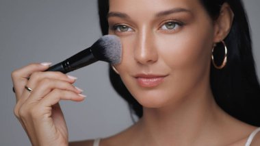 Siyah saçlı, halka küpeli, yüzünde makyaj fırçası tutan zarif bir kadının yakın çekimi. Kusursuz cildi ve parlak teni bu görüntüyü güzellik, cilt bakımı ve...