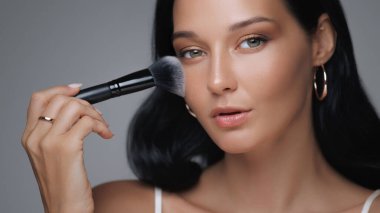 Göz kamaştırıcı genç bir kadın kusursuz cildini göstermek için dikkatlice bir fırçayla makyaj yapar. Güzellik, kozmetik, cilt bakımı ve yaşam tarzı promosyonları veya sosyal medya içeriği için mükemmel.