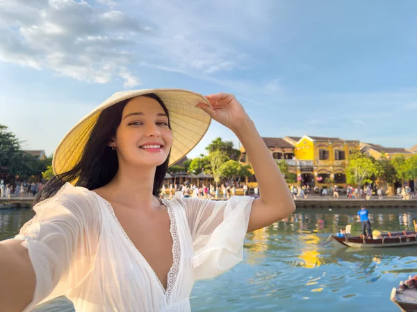 Vietnam 'da Hoi An nehrinin kenarında selfie çekerken koni şapkalı güzel bir kadın gülümsüyor. Parlak ve neşeli seyahat yaşam tarzı imajı reklamlar, tanıtımlar veya editoryal kullanım için mükemmel.