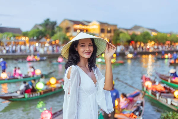 Geleneksel bir şapka ve beyaz elbise giyen çekici genç kadın Hoi An, Vietnam 'da fener ışığında yanan teknelerin önünde gülümsüyor. Seyahat yaşam tarzı, yayın ya da tanıtım içeriği için mükemmel.