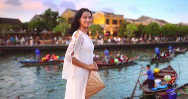 Akan beyaz elbiseli zarif Asyalı kadın Hoi An 'da nehir kenarında elinde konik bir şapka tutuyor. Gezi, moda ya da kültürel tanıtım için mükemmel..