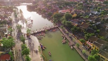 Hoi An 'ın çatılarında uçan ve teknelerle kalabalık bir nehir kenarı köprüsüne doğru yaklaşan, yatay formatdaki insansız hava aracı görüntüleri. Seyahat reklamları ve Asya yaşam tarzı projeleri için mükemmel..