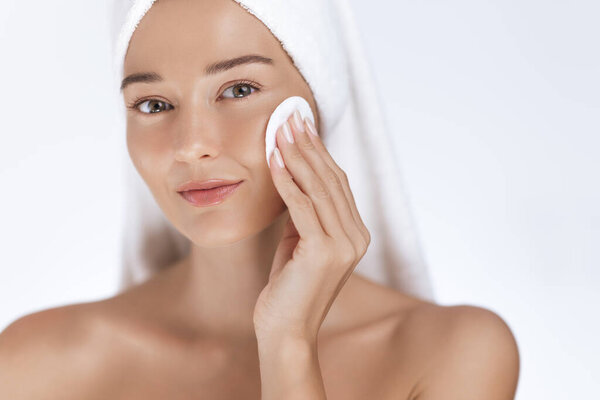 Caucasian woman with radiant skin uses a cotton pad to cleanse her face. Spa and beauty skincare concept with towel on head. Ideal for health, cosmetics, and wellness promotions.