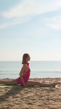 Pembe tek parçalı formalı genç bir kadın, deniz kenarındaki yoga minderinin ön tarafını tamamen ikiye ayırıyor. Sağlık, esneklik, fitness veya meditasyon temalı hisse senedi ve tanıtım videoları için ideal.