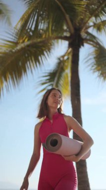 Pembe aktif giysili genç bir kadın tropik bir plajda palmiye ağaçlarının altında dikilirken yoga minderi tutuyor. Sağlık yaşam tarzı, yoga inzivası, fitness konsepti ve yüksek kaliteli hisse senedi için mükemmel.