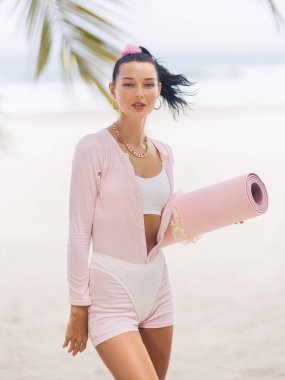 Güneşli bir plajda pembe yoga minderi tutan şık, genç, beyaz bir kadın. Şık giyimli, sağlıklı, spa, güzellik ve tropikal fitness içeriği için ideal..