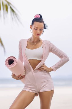 Pembe spor kıyafetleri içinde şık genç bir kadın kumlu tropik plajda yoga minderiyle poz veriyor. Modern nüfuzlu yaşam tarzı, sağlık ve moda konsepti, sosyal medya, reklamlar ve promosyon kullanımı için ideal