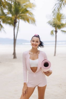Pembe elbiseli genç bir kadın gülümsüyor ve kum tropik plajda palmiye ağaçlarıyla yoga minderi taşıyor. Yüksek kaliteli yaşam tarzı ve sağlık konsepti, reklamlar, tanıtımlar, seyahat ve fitness için mükemmel