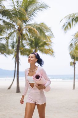 Elinde yoga minderi tutan ve palmiye ağaçlarıyla kumlu tropik plajda yürüyen zarif pembe elbiseli genç bir kadın. Yüksek kaliteli yaşam tarzı ve sağlık konsepti, reklamlar, tanıtımlar, seyahat ve fitness için mükemmel