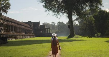 Angkor Wat tapınağı yakınlarında yürüyen geniş kenarlı şapkalı genç bir turist Kamboçya 'da harabe halinde. Yeşilliklerle çevrili tarihi Khmer mimarisi. Seyahat, kültür ve keşif konsepti görüntüleri.