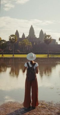 Sırt çantası ve şapkasıyla gölün kenarında durmuş Kamboçya 'daki Angkor Wat tapınağına bakan şık bir bayan gezgin. Seyahat reklamları, kültür mirası turizmi ve yaşam tarzı promosyonları için mükemmel stok görüntüleri..