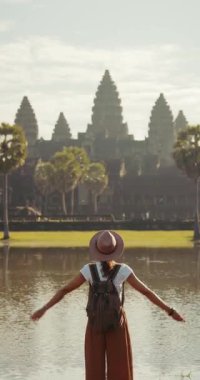 Kamboçya 'daki Angkor Wat tapınağının önünde el kaldıran sırt çantalı ve şapkalı neşeli kadın gezgin. Seyahat reklamları, yaşam tarzı turizmi, kültür mirası ve macera stoku tanıtımı için mükemmel bir görüntü..