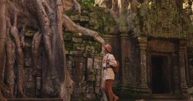 Şapkalı ve sırt çantalı genç bir kadın ünlü Ta Prohm Tapınağı 'nı keşfediyor. Büyük ağaç kökleri antik Khmer kalıntılarını kaplıyor. Seyahat blogları, kültürel belgeseller ve turizm için mükemmel stok videosu