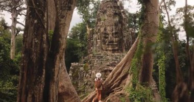 Sırt çantalı ve şapkalı kadın gezgin Zafer Kapısı yakınlarındaki dev ağaçların altında duruyor, Bayon Tapınağı, Angkor Thom, Kamboçya. Seyahat, kültür, turizm ve yaşam tarzı tanıtımı için yüksek kaliteli stok görüntüleri.