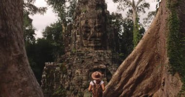 Devasa ağaç kökleri arasında duran şapkalı ve sırt çantalı kadın gezgin Zafer Kapısı, Bayon Tapınağı, Angkor Thom, Kamboçya. Turizm, yaşam tarzı ve kültürel tanıtımlar için yüksek kaliteli stok videosu.