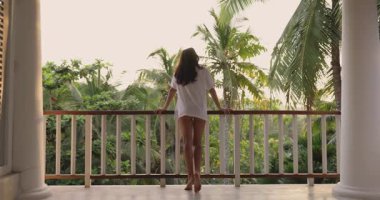 Gevşek beyaz gömlekli genç bir kadın, nefes kesici tropikal manzarayı izlerken gülümseyerek balkona yaslanıyor. Huzurlu ortam rahatlamayı, mutluluğu ve doğanın güzelliğini vurgular..