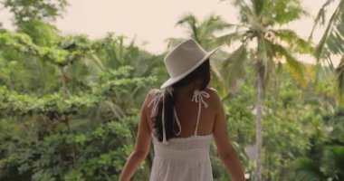 Beyaz yaz elbisesi ve şapkası giyen şık genç bir kadın. Tropikal tatil köyünün balkonunda, yemyeşil ormana bakan. Mükemmel yüksek kaliteli yaşam tarzı, seyahat, tatil ve sağlık stoku video konsepti.