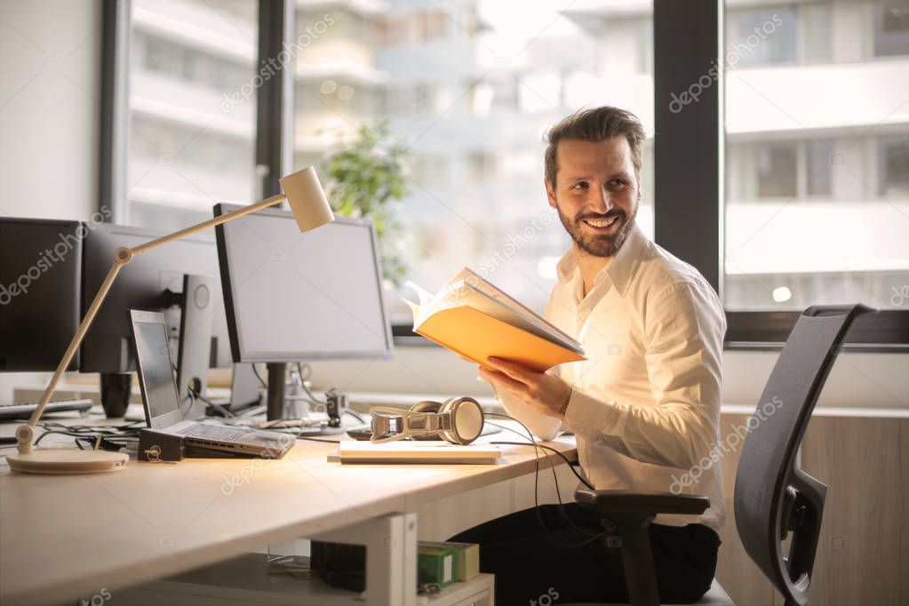 Young charming business man sitting at his work desk, holding a book and smiling. 