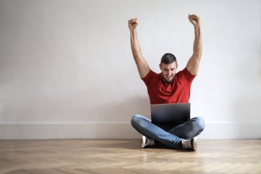 Kapalı yere oturmak, onun laptop kontrol ve tamamen mutlu duygu genç adam.